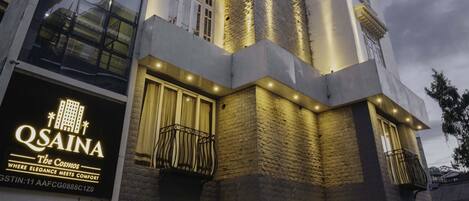 A modern hotel with a prominent 'QSAINA' sign, large windows, and a well-lit entrance.