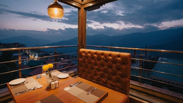 A dining table set with plates, cutlery, and a vase of yellow flowers, overlooking a mountainous landscape. - Q Saina The Cosmos (Gangtok)