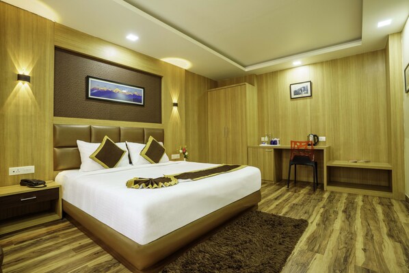 A hotel room with a large bed, wooden panel walls, a desk with a chair, and a small table. - Q Saina The Cosmos (Gangtok)