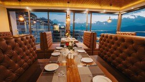 A dining area with a wooden table set for four, surrounded by leather booths, with a mountain view through large windows. - Q Saina The Cosmos (Gangtok)