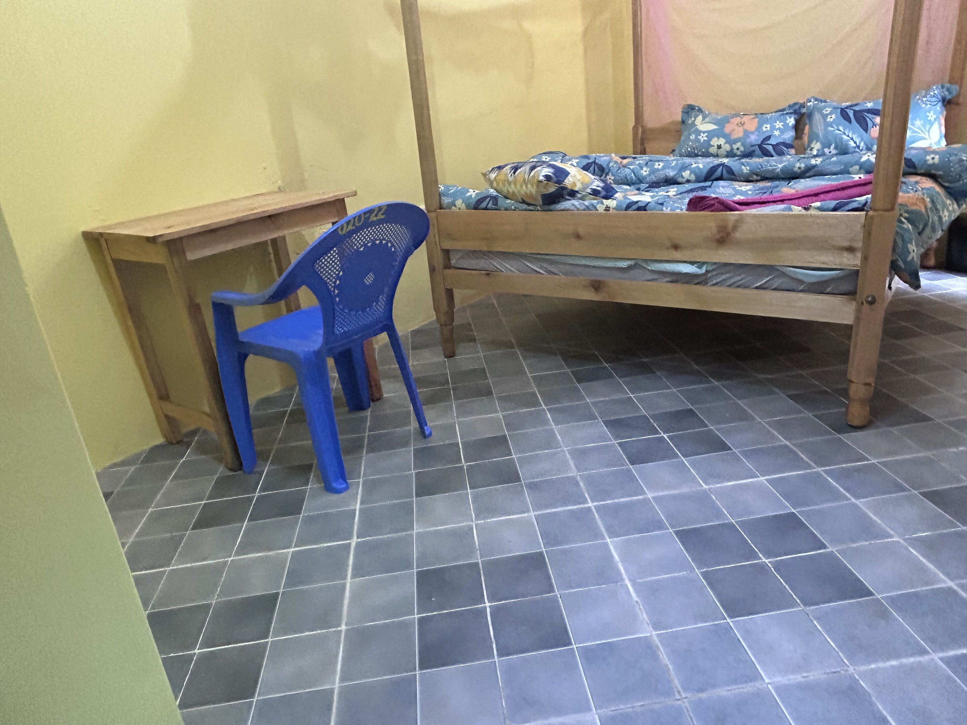 A room with a bed, a blue plastic chair, a small wooden desk, and a tiled floor.