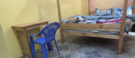 A room with a bed, a blue plastic chair, a small wooden desk, and a tiled floor.