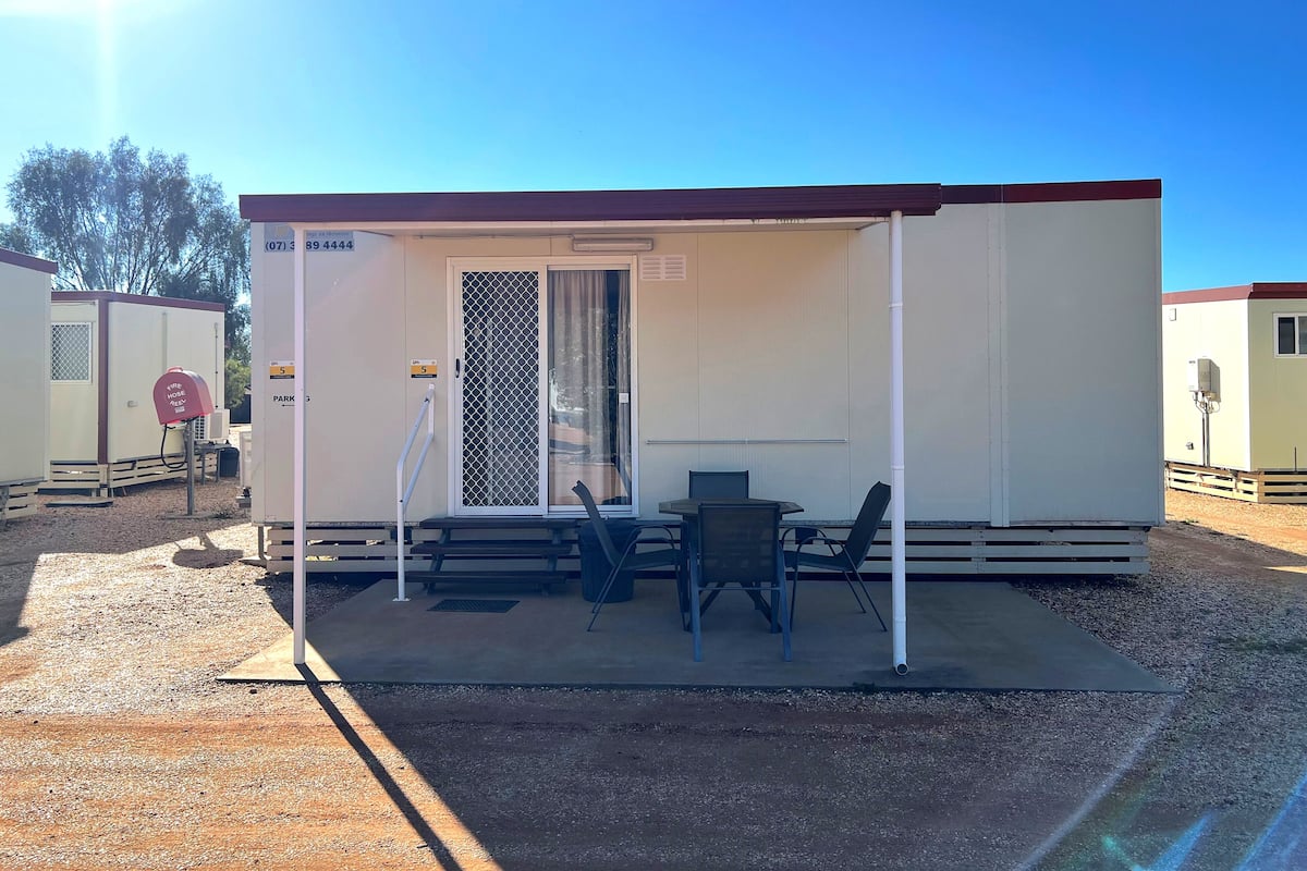 A small, single-story mobile home with a covered porch, a table and chairs, and a fire hose access point.