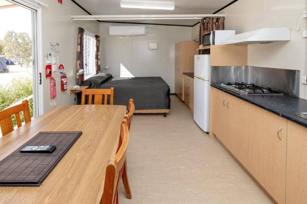 A compact kitchen with a stove, microwave, and refrigerator. There is a bed with a gray mattress and a window with curtains. A table with chairs is positioned near the window.