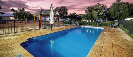 A rectangular swimming pool with a wooden deck, surrounded by a fence, and a thatched-roof structure.