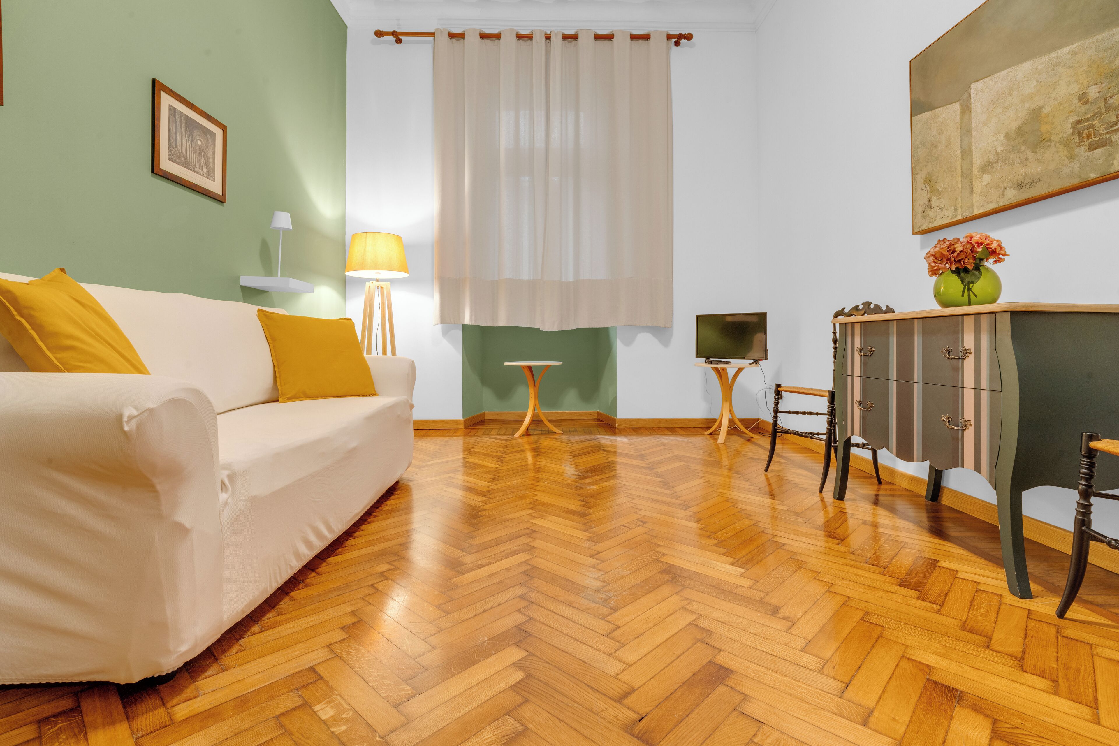 A room with a wooden herringbone floor, a white sofa with yellow cushions, a small table with a lamp, a cabinet with a vase of flowers, and a painting on the wall.