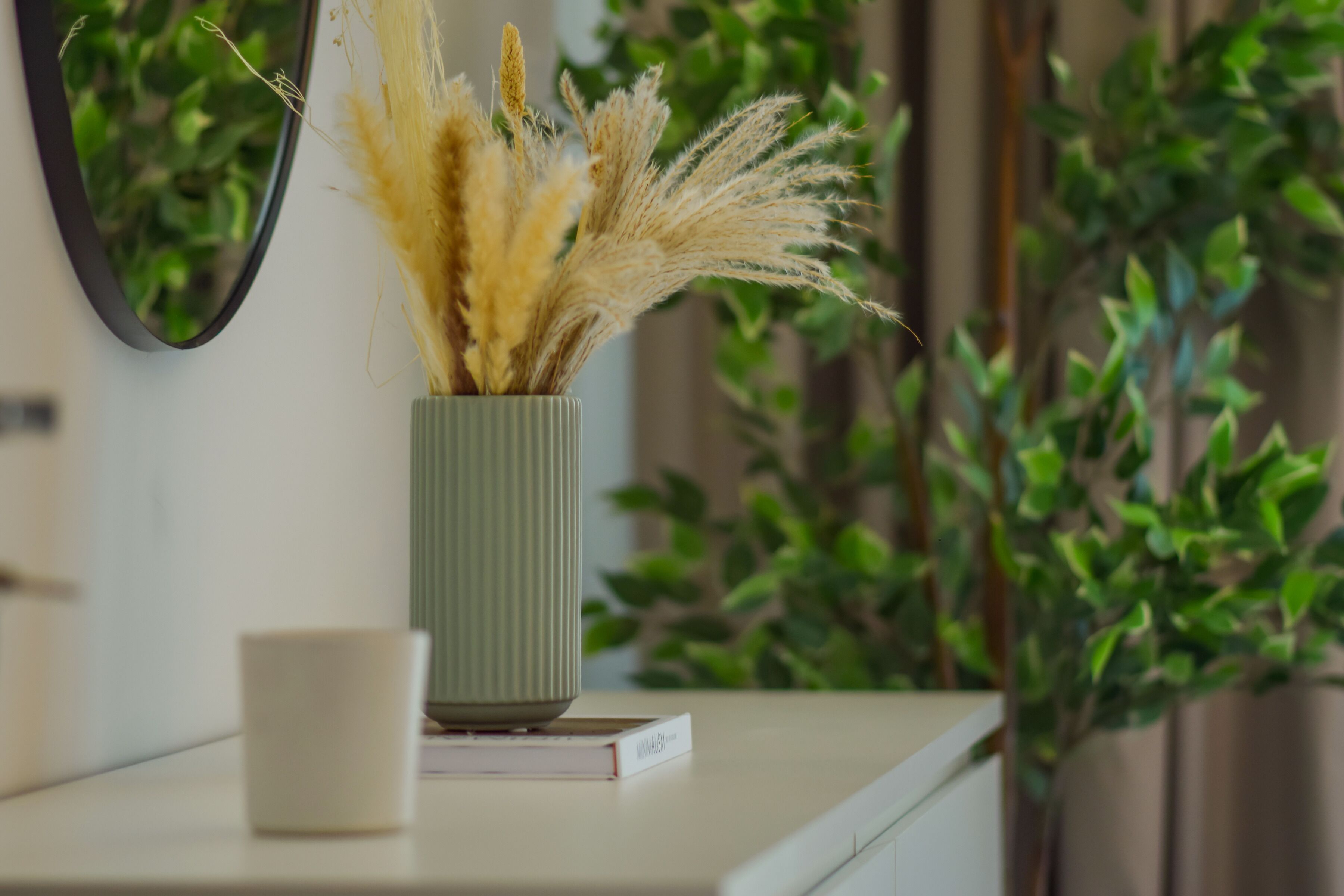 A vase with dried plants on a white surface, a mirror, and a green plant in the background.