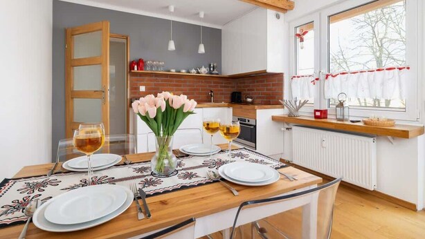 A modern kitchen with a wooden table set for dining, a vase with flowers, and a window with curtains.