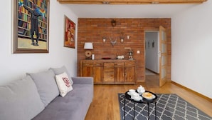 A living room with a grey sofa, a black and white patterned ottoman, a wooden coffee table with teapots and cups, a brick wall with mounted animal heads, and a bookshelf with a colorful painting.