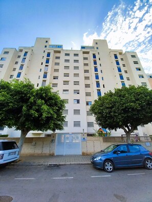 Un edificio residencial de varios pisos con un acceso cerrado, un coche azul estacionado al frente y árboles en primer plano.