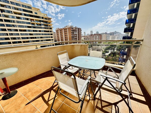 Un balcón con una mesa y sillas, una silla con cojín, y vistas de edificios cercanos.
