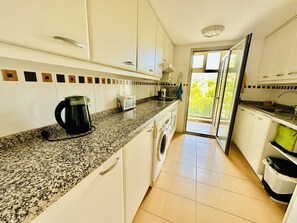 A kitchen with white cabinets, a granite countertop, a coffee maker, a washing machine, and a view of greenery outside.