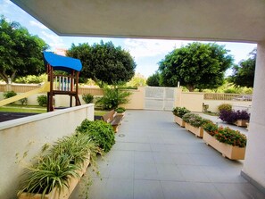 A covered outdoor area with a playground, benches, potted plants, and a gated entrance.