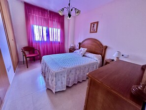A bedroom with a bed, a chair, a desk, a nightstand, a lamp, and a window with curtains.