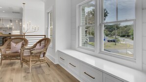 Une pièce dotée d’une grande fenêtre, de chaises en osier, d’un banc blanc et, au fond, d’une cuisine moderne.