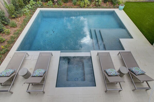 Une piscine rectangulaire avec un jacuzzi au centre, entourée de chaises longues dotées de coussins à motifs.