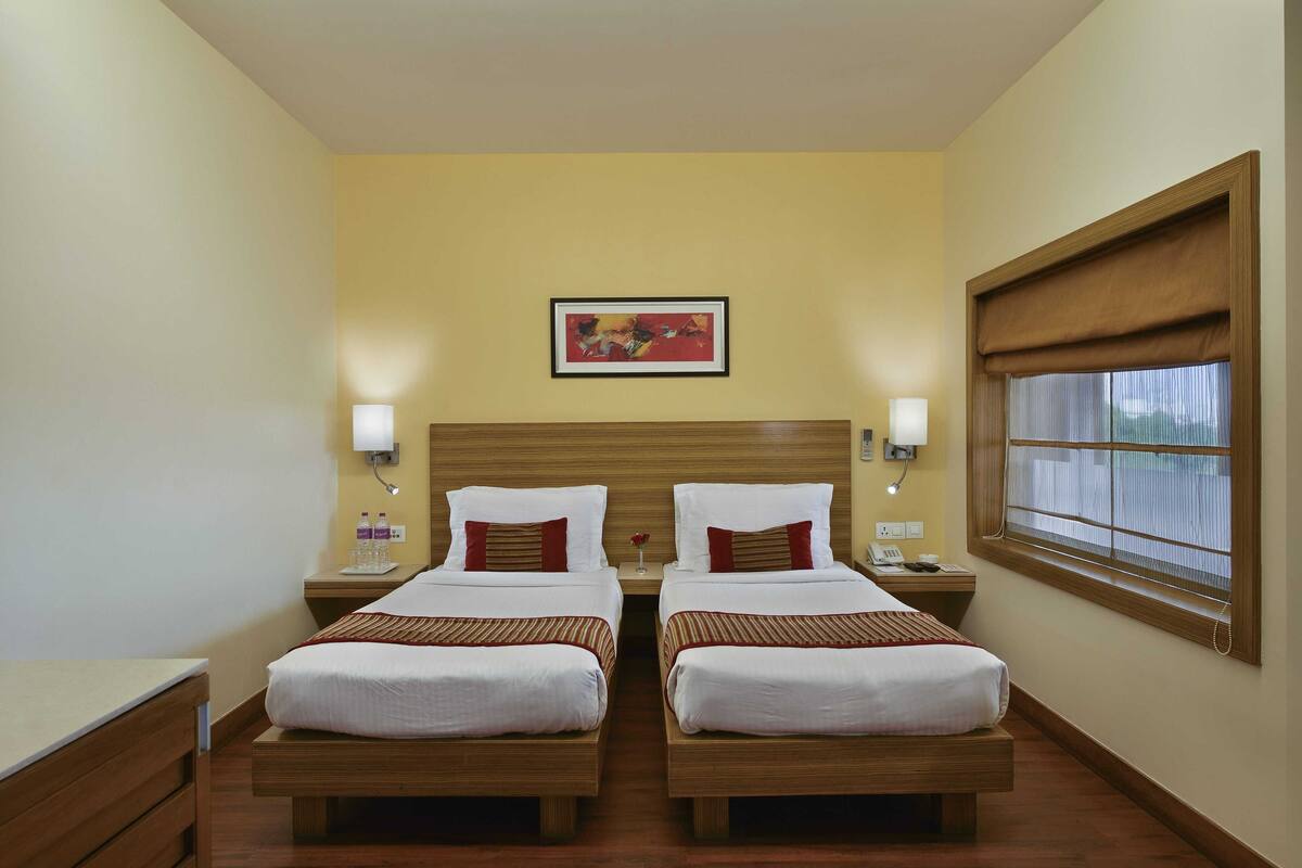A hotel room with two beds, a wooden headboard, bedside lamps, a window with a blind, and a framed picture on the wall.