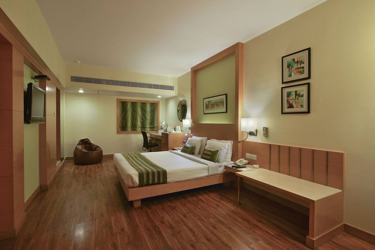 A hotel room with a wooden floor, a bed with a green and white bedspread, a desk with a telephone, a chair, a small round table, and a green curtain.