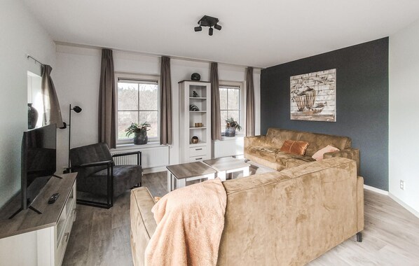 Un salon moderne avec un canapé beige, une table basse et un téléviseur. De grandes fenêtres sont agrémentées de rideaux, et on y trouve une bibliothèque ainsi qu'un tableau accroché au mur.