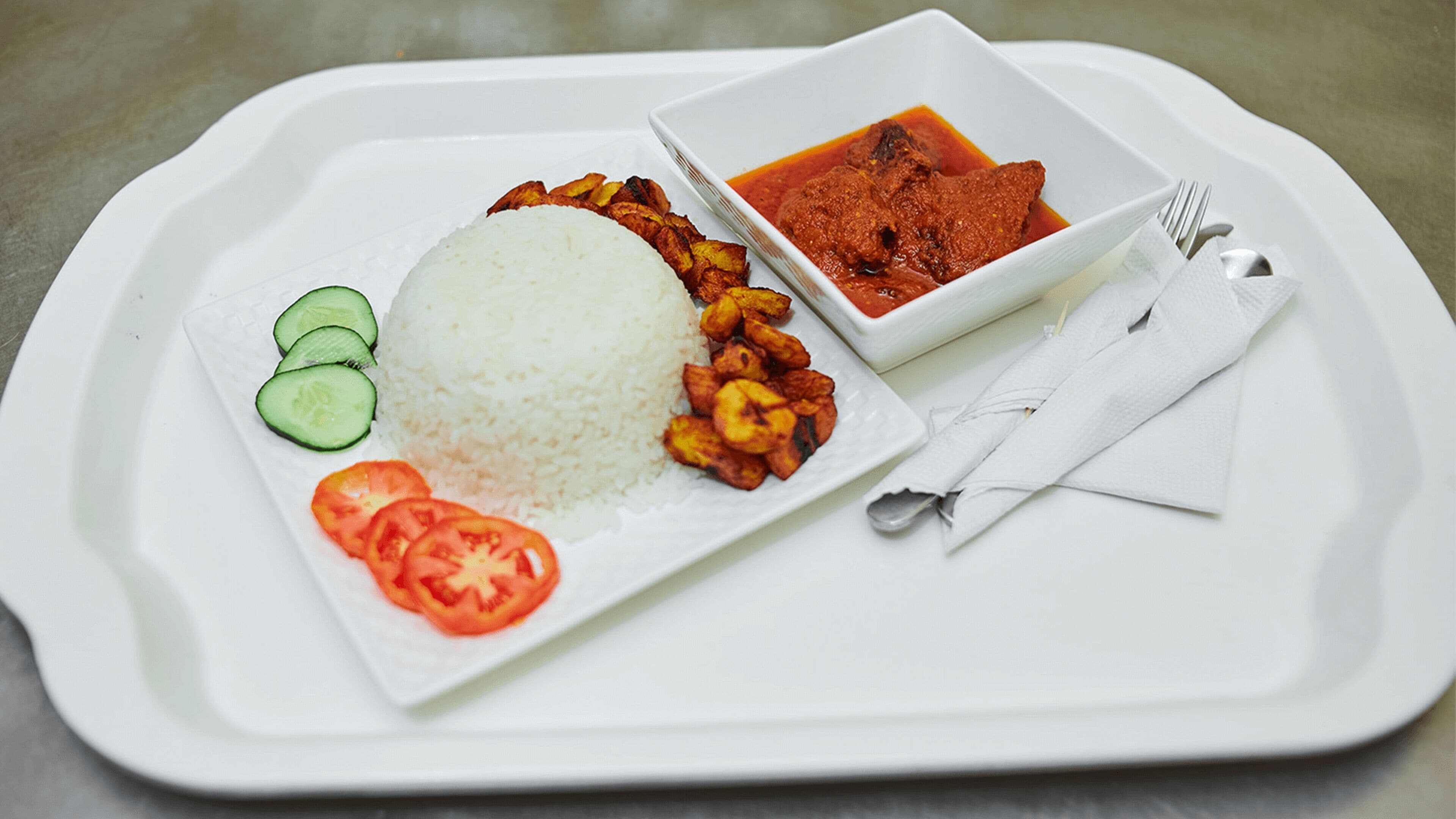 A tray with white rice, sliced tomatoes, cucumber slices, fried plantains, and a bowl of red curry.