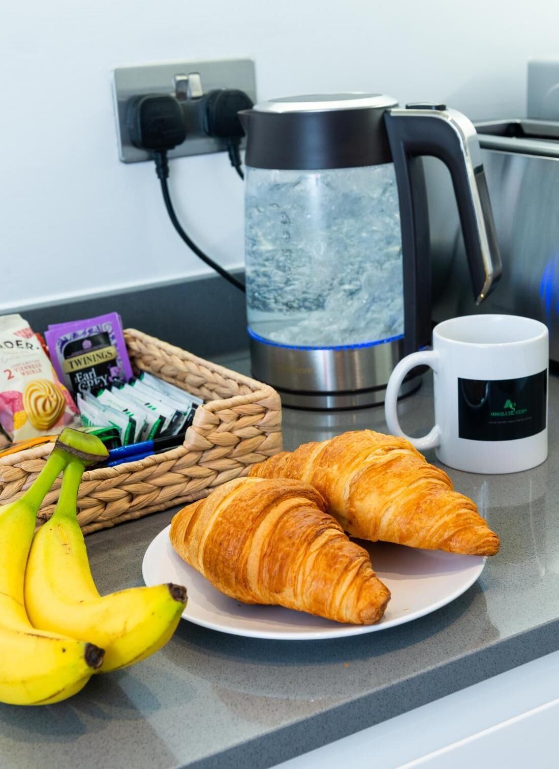 A breakfast setting with croissants, bananas, a coffee maker, and a mug on a kitchen counter.