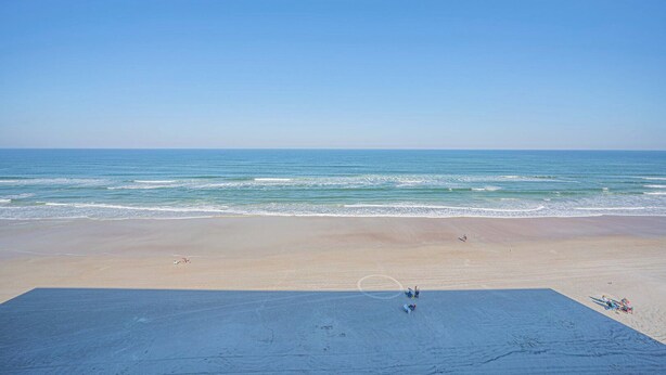 Ein Strand mit blauem Himmel und ruhiger See. Menschen und Liegestühle liegen im Sand.