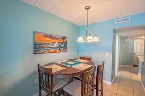A dining area with a round table, chairs, a painting of a sunset over the ocean, and a ceiling light fixture.