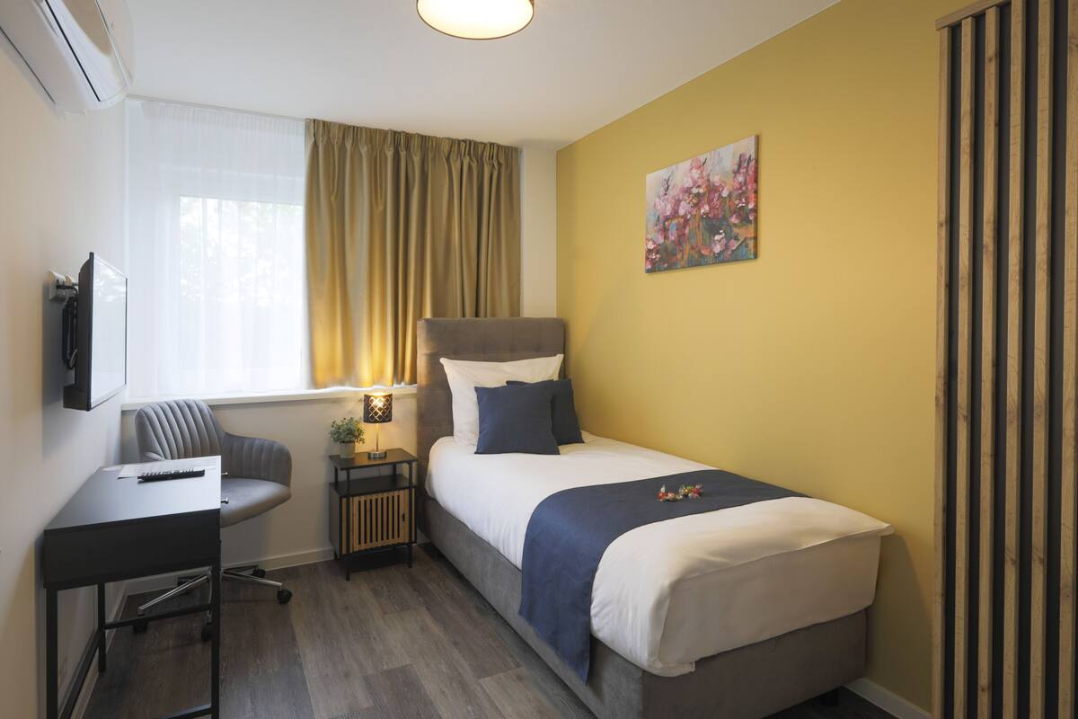 Habitación de hotel con cama, escritorio, silla, mesita de noche con lámpara y ventana con cortinas.