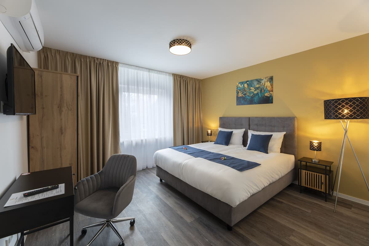 Habitación de hotel con cama, escritorio, silla, lámpara y un cuadro en la pared.