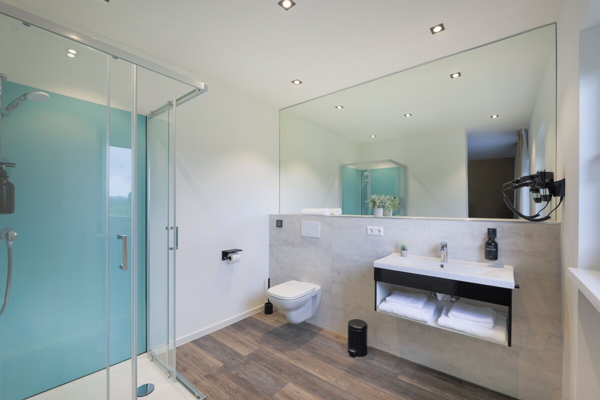 A modern bathroom with a glass shower enclosure, a white toilet, a sink with a mirror, and a black trash bin.