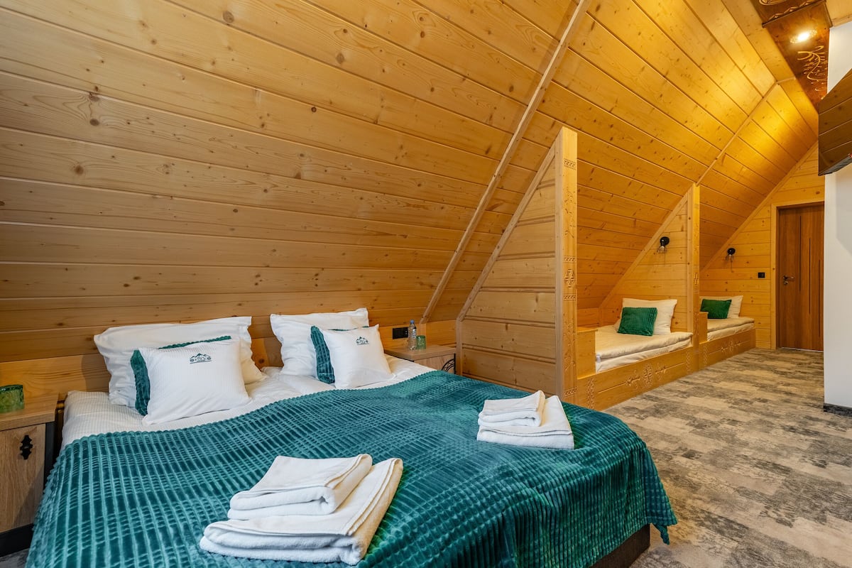 A cozy wooden cabin bedroom with a loft bed, a green bedspread, and neatly folded towels on the floor.