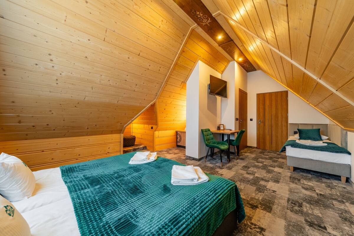 A cozy attic bedroom with a sloped ceiling, wooden walls, a bed with a green blanket, a desk, and a chair.
