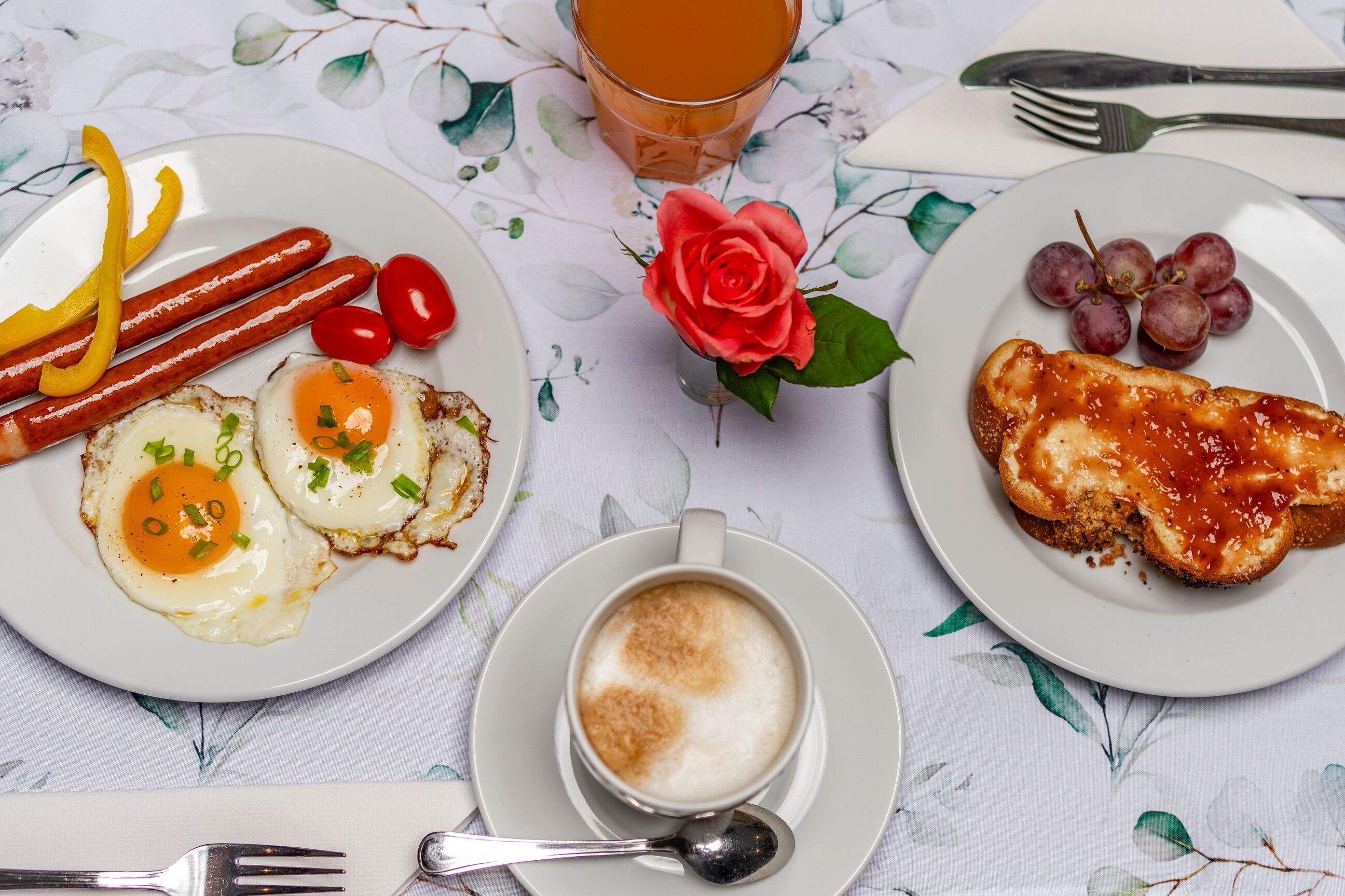 Un desayuno con platos de comida, una taza de café y un vaso de zumo de naranja sobre una mesa con mantel floreado.