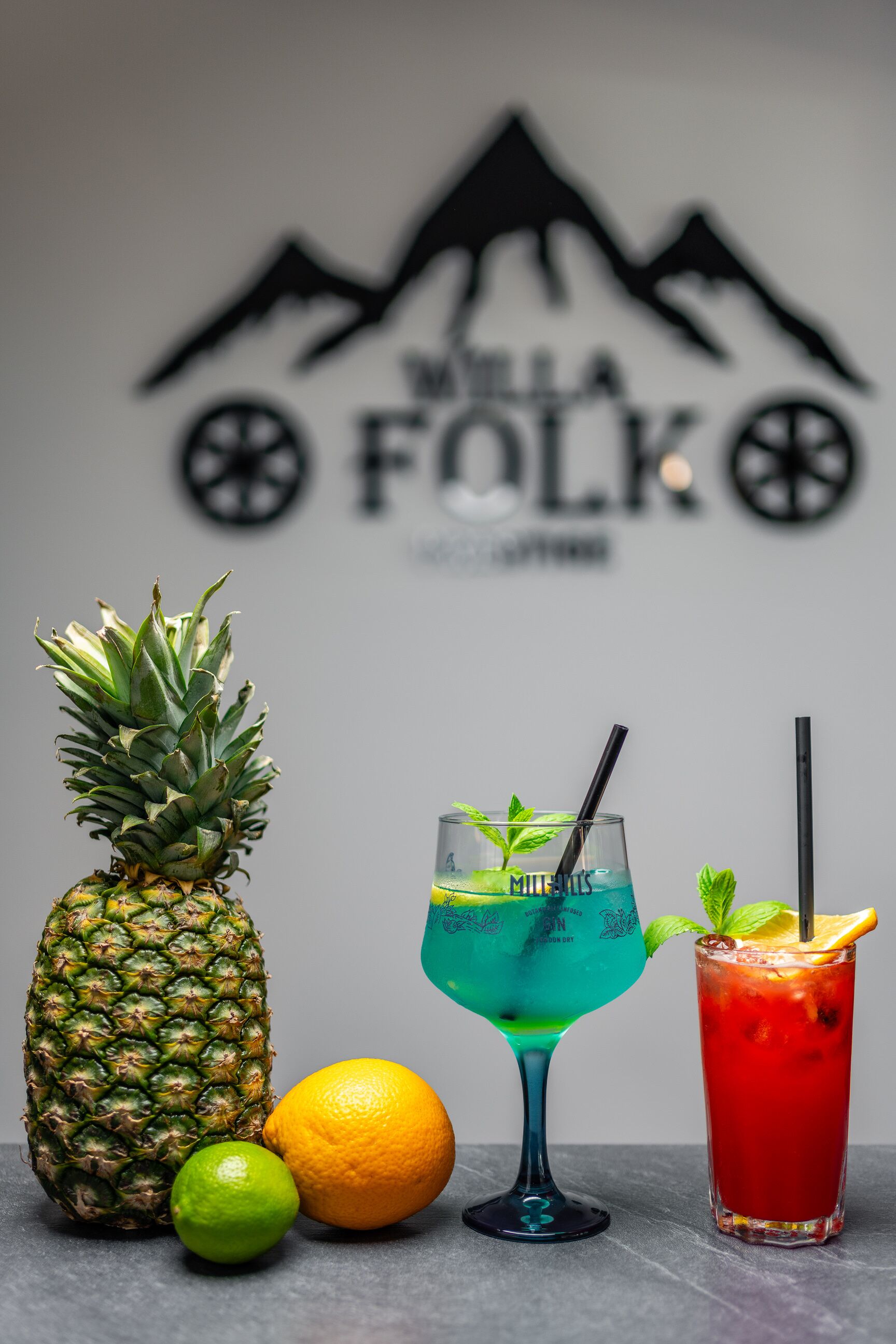 Una piña, un limón, una naranja y dos bebidas sobre una mesa, con un logotipo al fondo.