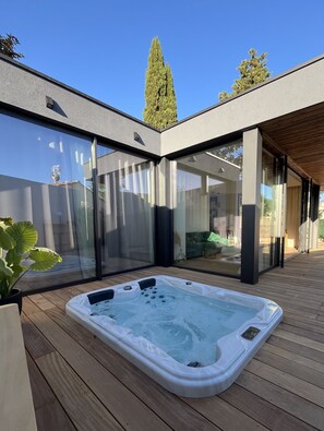 A modern outdoor hot tub area with a wooden deck, large sliding glass doors, and a tall tree in the background. - Lodge Contemporain Avec Jacuzzi en Provence Verte (Tourves)