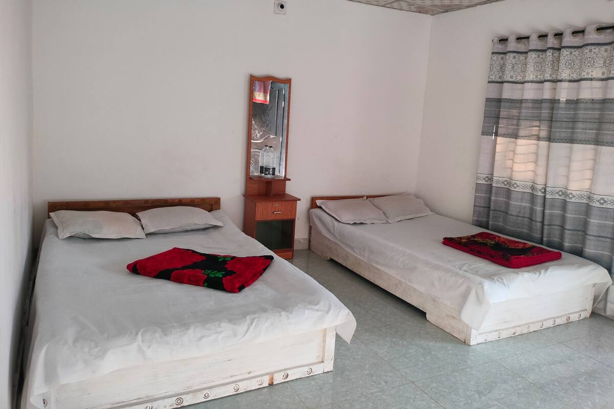 A room with two single beds, a wooden nightstand with a mirror, a ceiling fan, and a window with patterned curtains.