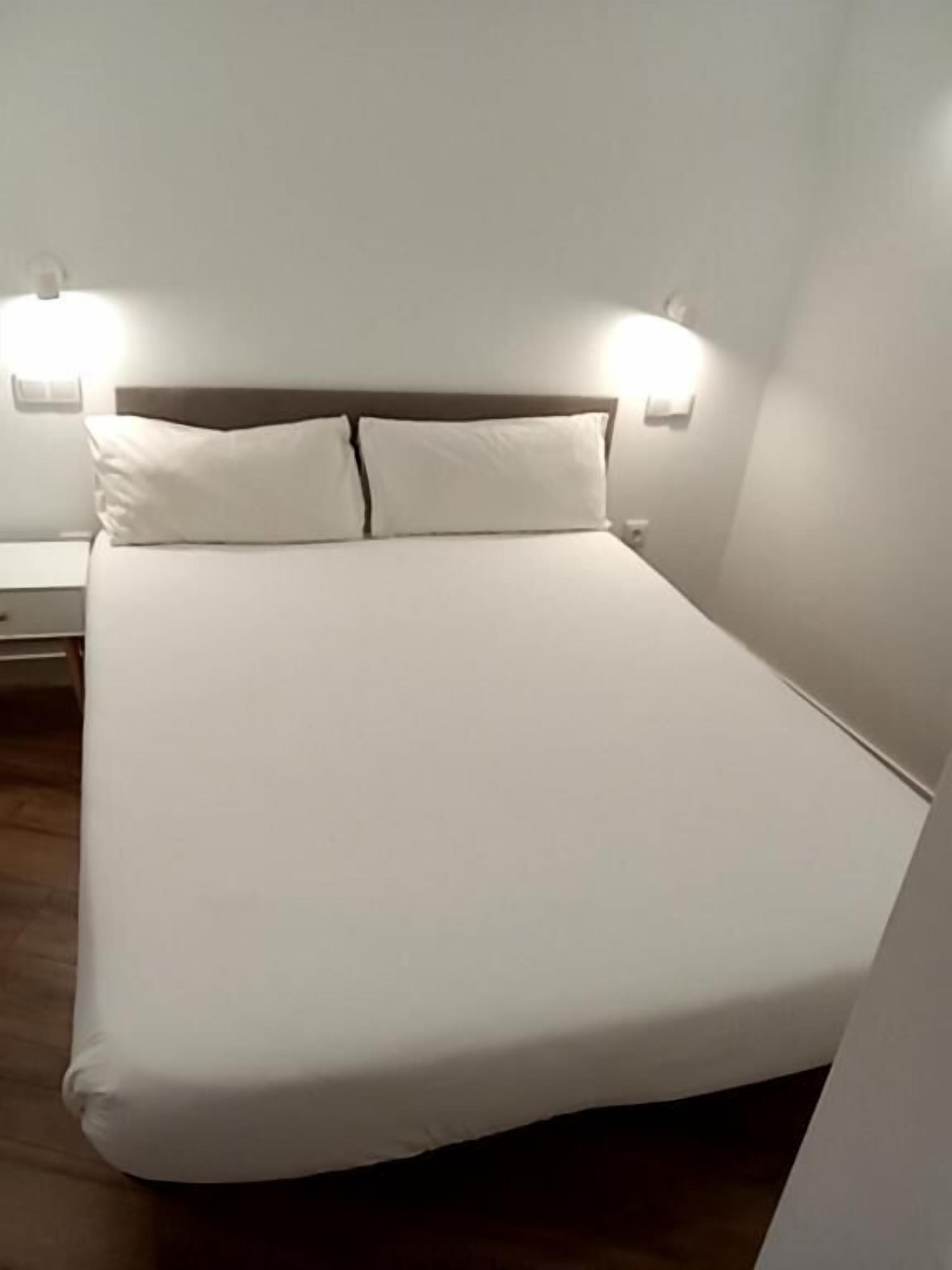 A neatly made bed with white linens and a wooden headboard. There are two bedside tables with lamps on either side of the bed.