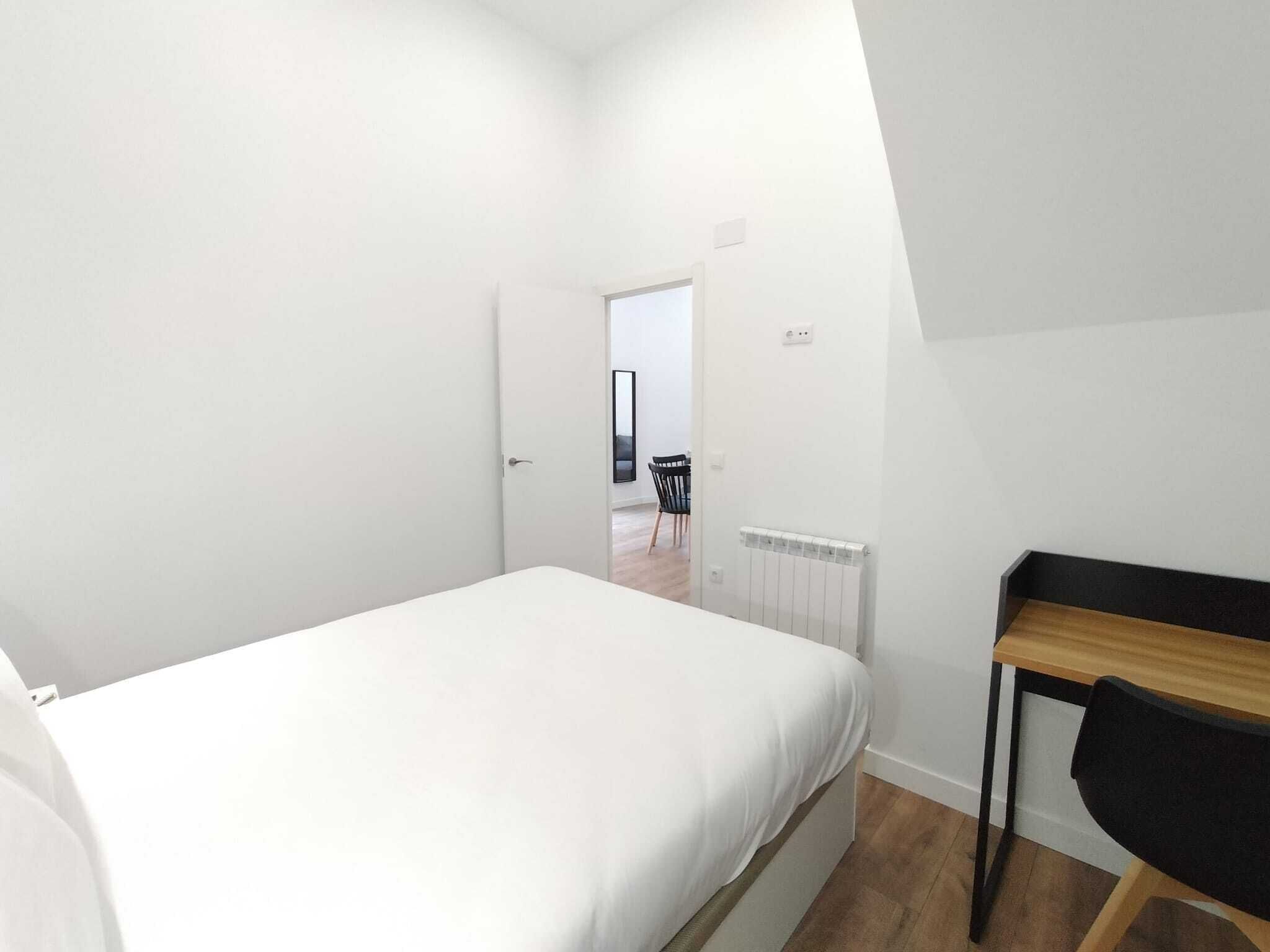 Une chambre à coucher avec un lit, un bureau et une chaise. Il y a un radiateur au mur et une porte qui donne sur une autre pièce.