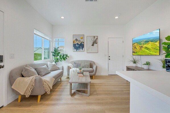 Un salon spacieux avec un canapé, une table basse et un téléviseur. Deux tableaux ornent le mur et une grande fenêtre laisse entrer la lumière naturelle.