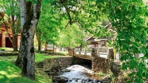 Un puente de piedra sobre un arroyo serpenteante en un parque con árboles y un edificio tradicional al fondo.