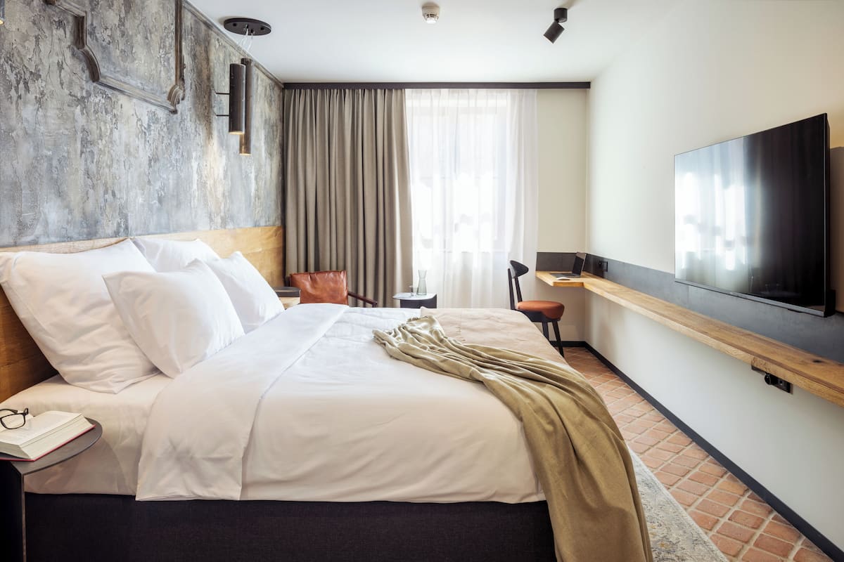Ein modernes Hotelzimmer mit einem großen Bett, einem Flachbildfernseher, einem Holfnachtisch, einem Sessel und einem großen Fenster mit Vorhängen.