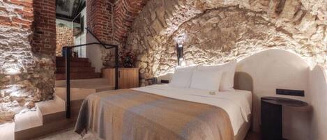 A bedroom with a stone wall, a bed, a bedside table, and a staircase leading to an upper level.