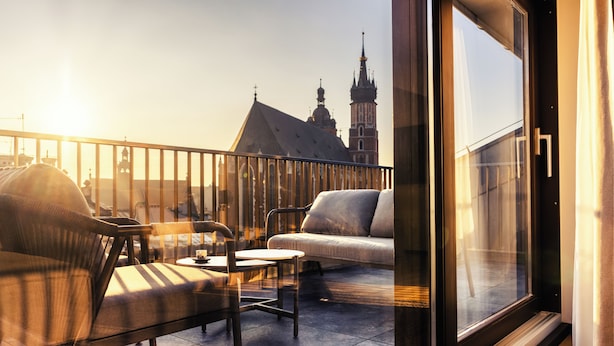 A balcony with a sofa, armchair, and table, overlooking a building with a tower. - Nomadic Urban Premium Suites (Kraków)