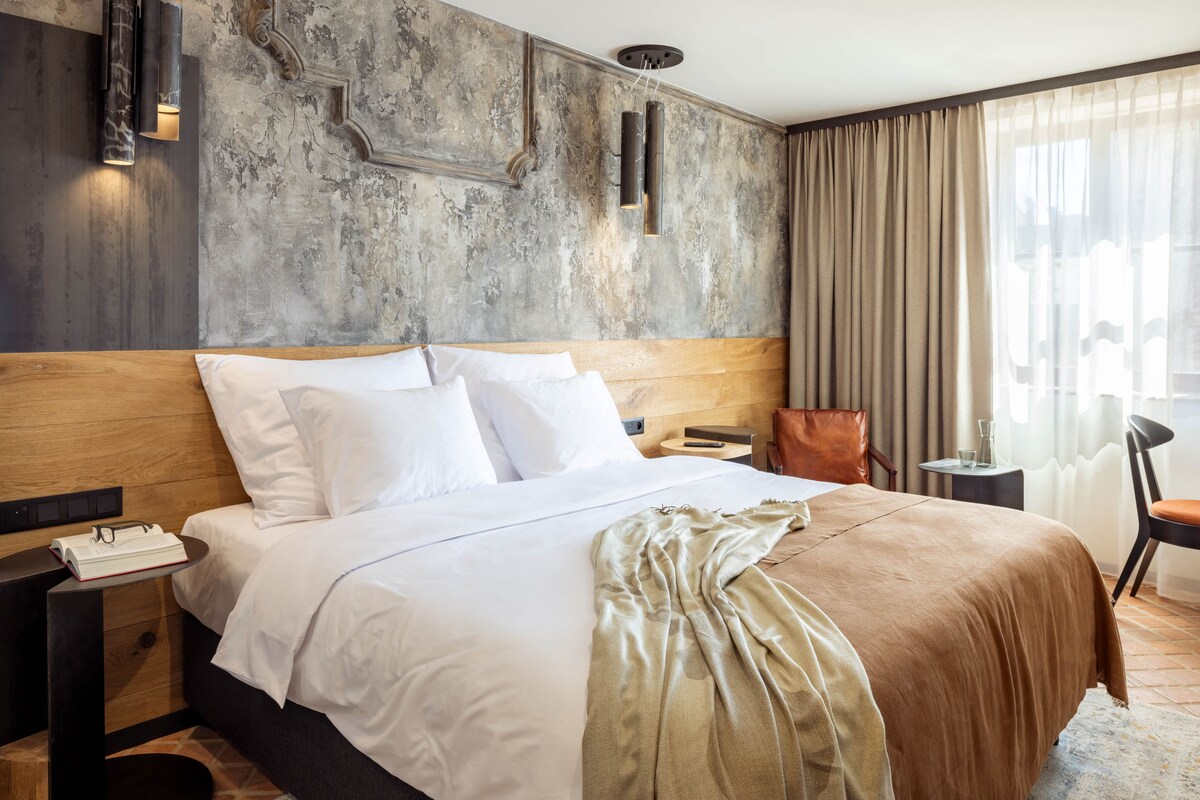 A hotel room with a large bed, a wooden headboard, a nightstand with a lamp, a chair, and a view of the city through sheer curtains.