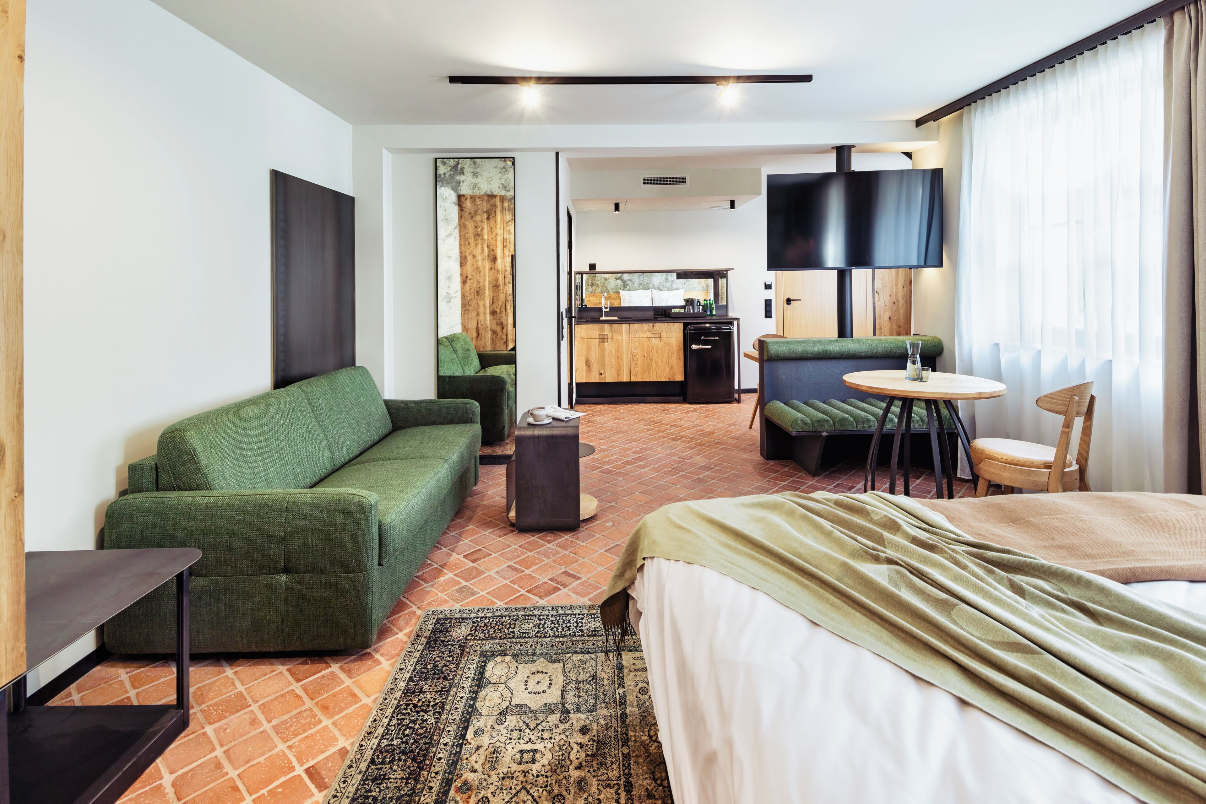 Ein Hotelzimmer mit einem grünen Sofa, einem Bett mit grüner Decke, einem kleinen runden Tisch, einem Sessel, einem Fernseher und einer Küchenzeile im Hintergrund.
