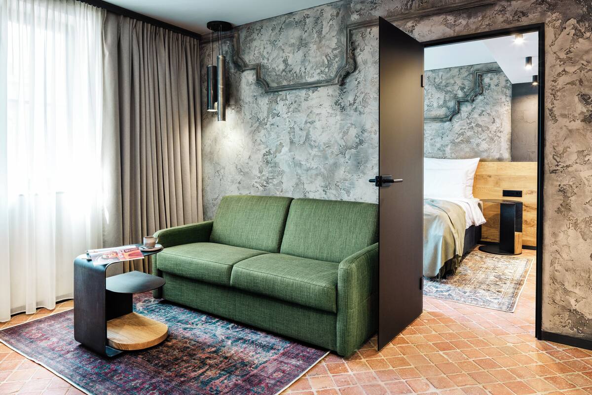 Ein modernes Zimmer mit einem grünen Sofa, einem kleinen Beistelltisch, einem gemusterten Teppich und einem Bett mit Holz Kopfteil.