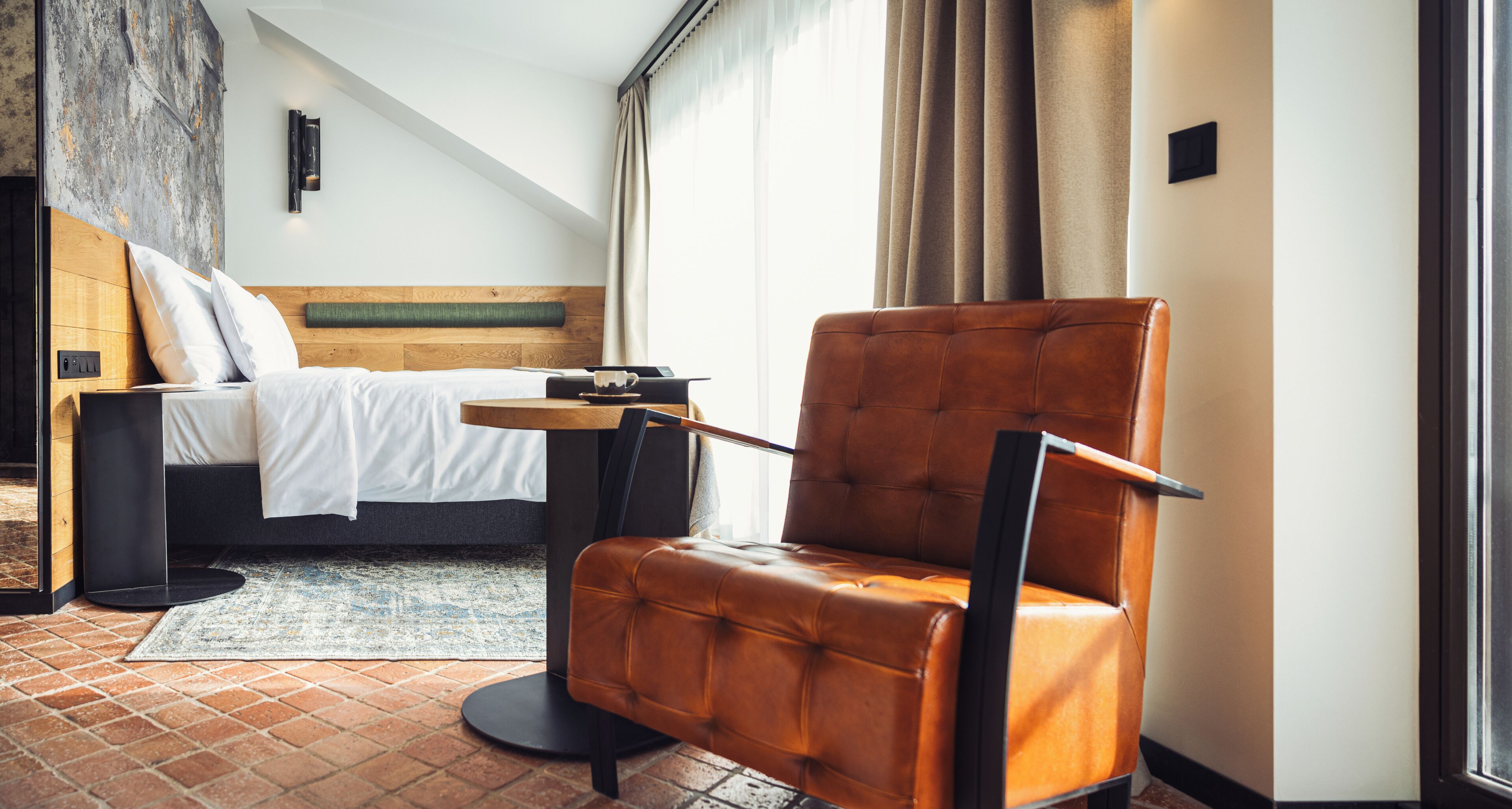 A modern hotel room with a bed, a leather armchair, a small table, and a view of the outdoors.