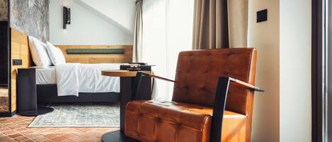 A modern hotel room with a bed, a leather armchair, a small table, and a view of the outdoors.