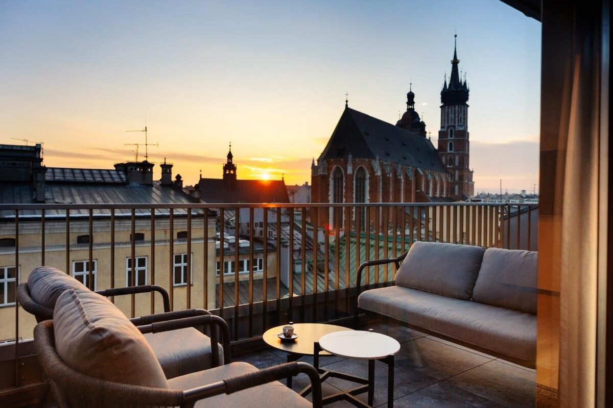 A balcony with a sofa, a chair, and a small table, overlooking a historic building with a church and a tall tower.