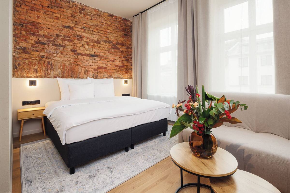 Ein Hotelzimmer mit Backsteinwand, einem Bett, einem Sofa, einem kleinen Tisch mit Blumenvase und einem fenster mit Vorhängen.